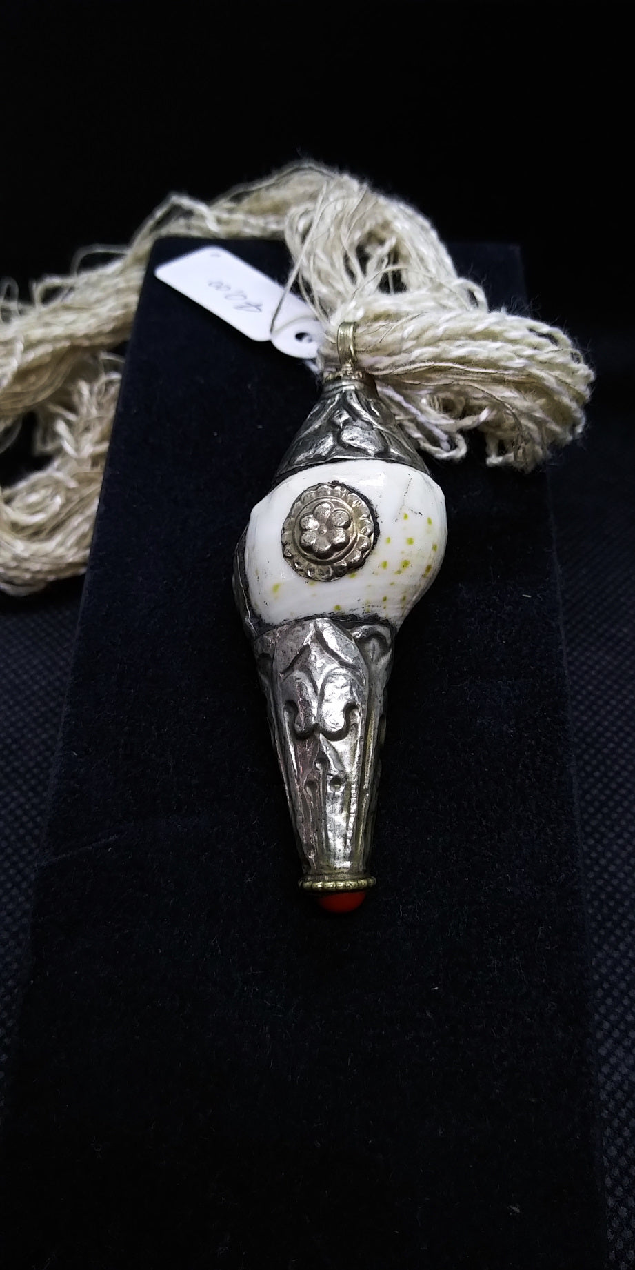 pendaglio raffigurante una conchiglia tibetana con argento cesellato e corallo . oggetto di culto di cultura buddista , strumento rituale. Era già utilizzata in epoca prebuddhista come simbolo delle divinità femminili, come contenitore e come strumento musicale rituale. Nel Buddhismo tibetano si utilizza spesso come strumento musicale e il suo potente suono viene utilizzato per richiamare i monaci alle riunioni, per fare offerte di suono durante le puje. Rappresenta la gloria dell’insegnamento del Dharma, c