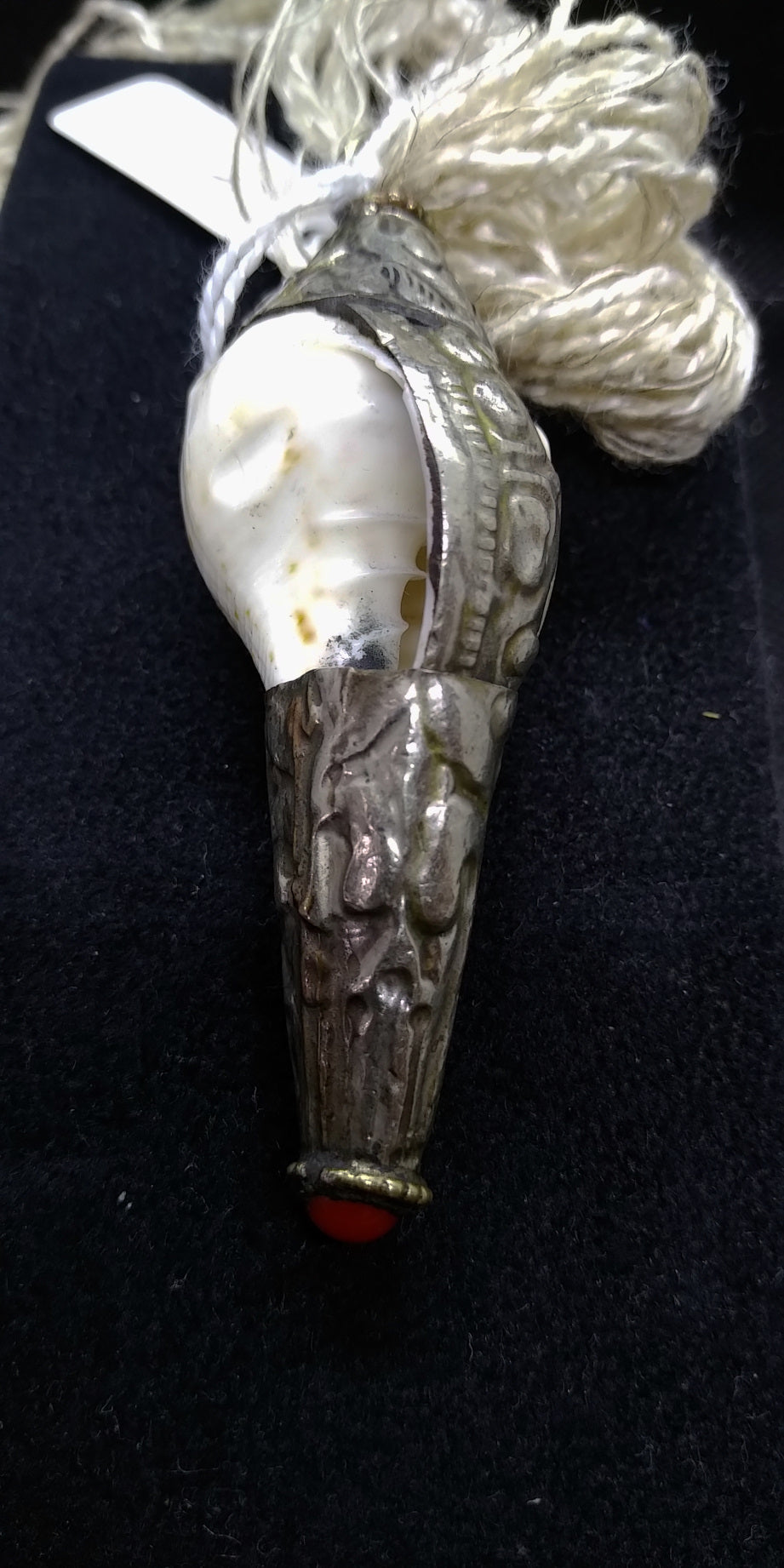 pendaglio raffigurante una conchiglia tibetana con argento cesellato e corallo . oggetto di culto di cultura buddista , strumento rituale. Era già utilizzata in epoca prebuddhista come simbolo delle divinità femminili, come contenitore e come strumento musicale rituale. Nel Buddhismo tibetano si utilizza spesso come strumento musicale e il suo potente suono viene utilizzato per richiamare i monaci alle riunioni, per fare offerte di suono durante le puje. Rappresenta la gloria dell’insegnamento del Dharma, c